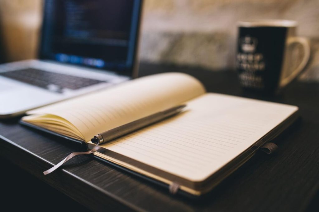 open notebook and pen next to a laptop on a wooden desk, daylight, no people